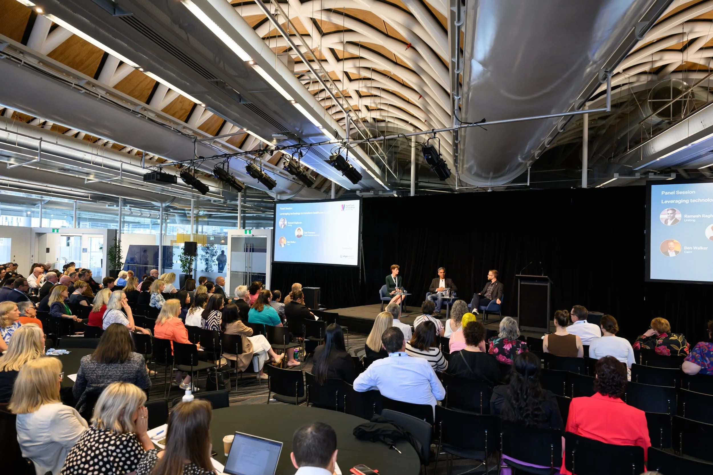 ACA conference panel discussion taking place in a large, modern auditorium with many attendees seated and listening to the panelists on stage. The stage has three speakers, two women and one man. Ben Walker is facilitating the panel.