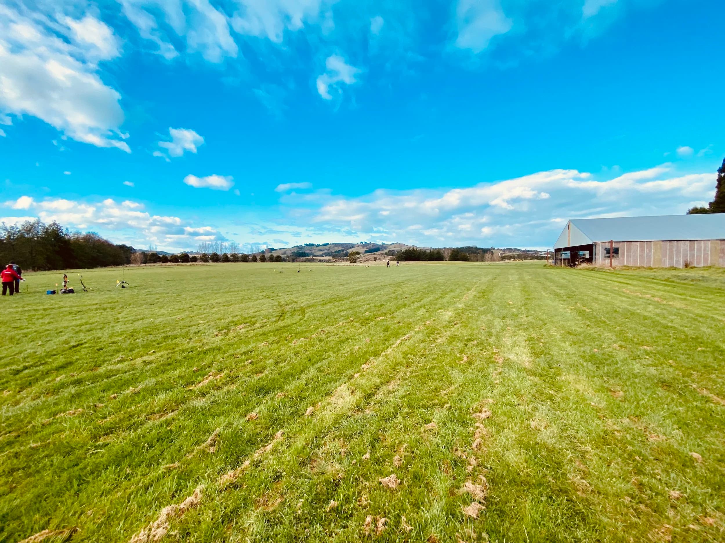 Wide green field with people on the left, a large barn on the right, blue sky with clouds, and distant hills.