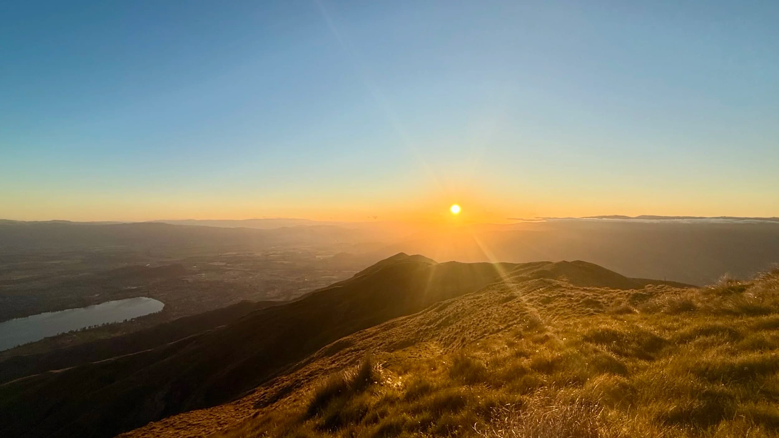 Sunset over rolling green New Zealand hills with a lake in the distance.