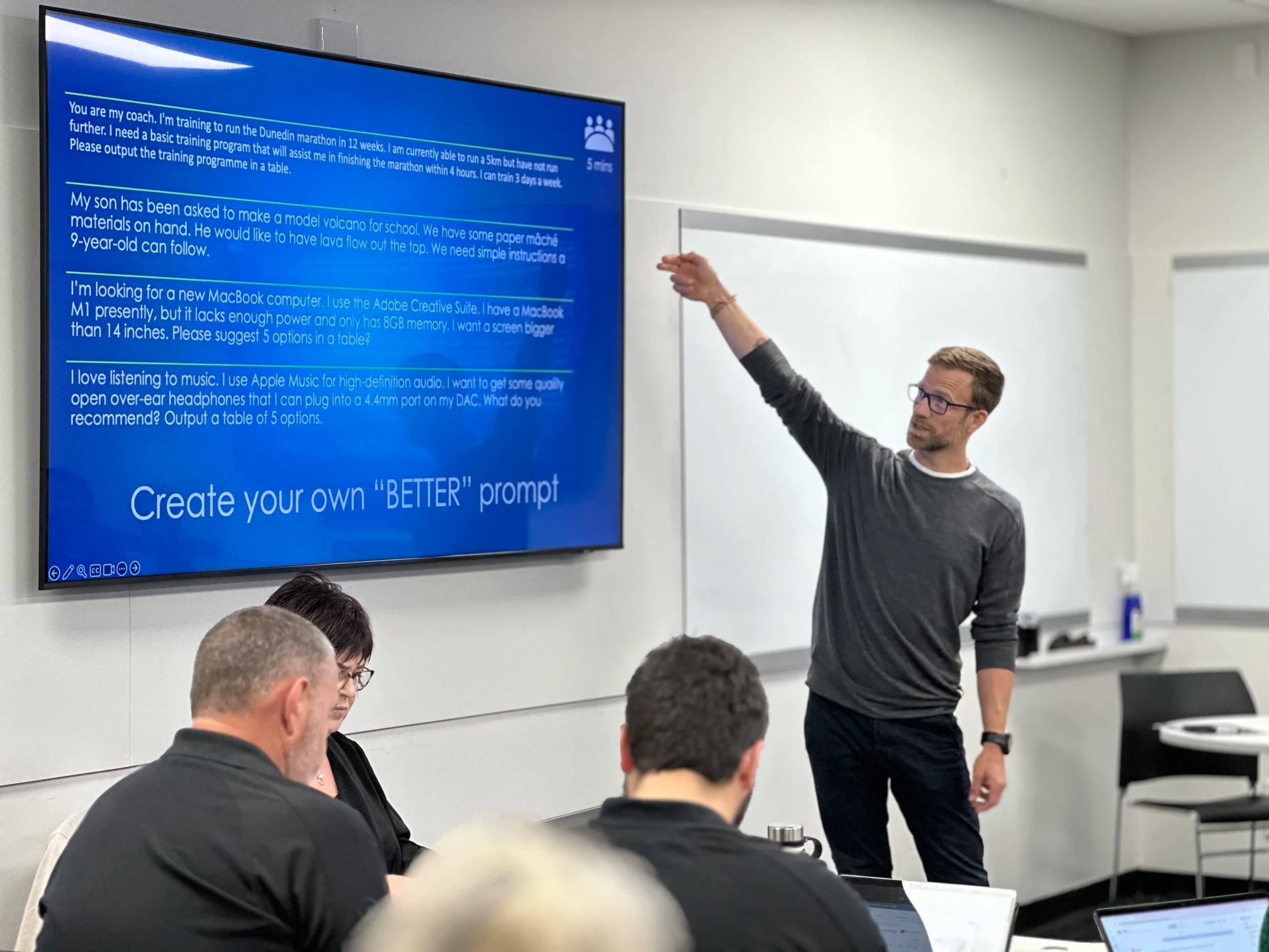 Ben Walker in glasses and a gray sweater is facilitating an AI workshop in a classroom, pointing at a large screen displaying a list of user prompts and responses. Several students are seated, viewing laptops.
