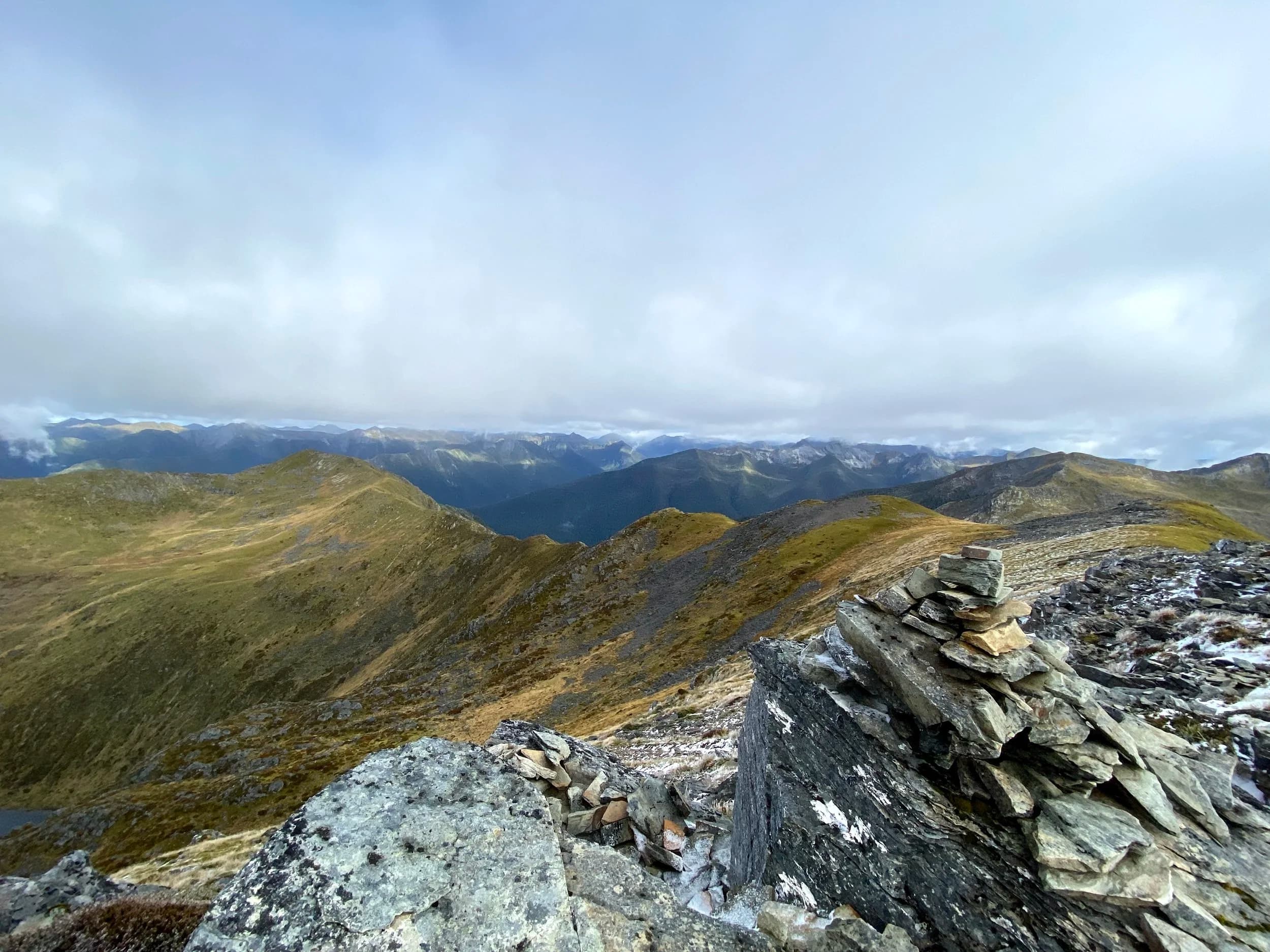 Mountain landscape with grassy ridges and distant peaks under cloudy sky, with a cairn of stacked stones in the foreground.